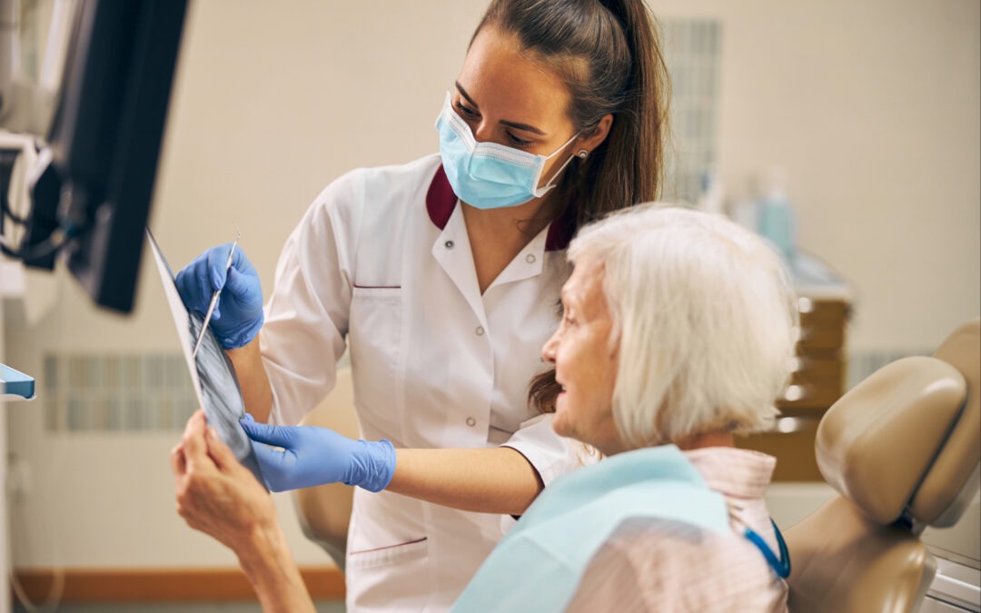 Zahnärztin mit Mundschutz und Handschuhen erklärt einer älteren Patientin im Behandlungsstuhl anhand eines Röntgenbilds die Zahnsituation in einer modernen Zahnarztpraxis.
