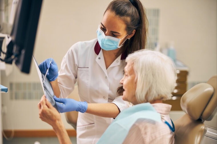 Zahnärztin mit Mundschutz und Handschuhen erklärt einer älteren Patientin im Behandlungsstuhl anhand eines Röntgenbilds die Zahnsituation in einer modernen Zahnarztpraxis.